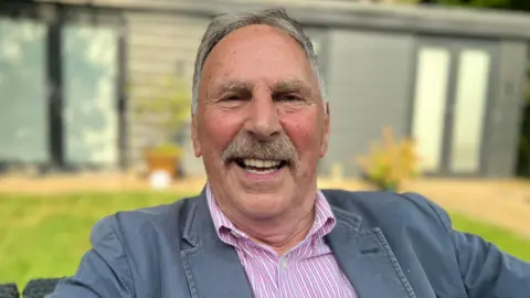 Emma Baugh/BBC Present day photo of Mr Dowson who is smiling joyfully at the camera. He has grey hair, a moustache and wears and pink and white striped shirt with a grey blazer style jacket over the top. The background of the image is blurred but it is noticeable that he is sat outside. Behind him, green grass can be made out as well as a grey panelled form of outhouse.  