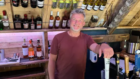 A man is stood with his hand on a beer tap while stood behind a bar looking at the camera.