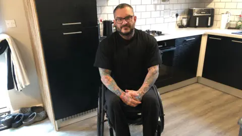 Darren Rozier/BBC Mr Hewitt is pictured in his wheelchair in the kitchen in his home. He is wearing a black t-shirt and black jogging bottoms. His hands are placed one in the other in his lap. There are tattoos along the length of both of his arms.