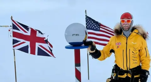 A man in a yellow top and red sunglasses and gloves in a polar setting with a union flag and stars and stripes in the background.