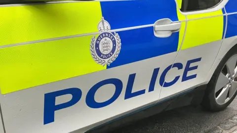 BBC The side of a Guernsey police car with the Guernsey Police crest in the centre of the car door. The car is coloured white with neon yellow and blue coloured livery.