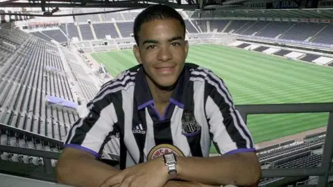 PA Media Kieron Dyer wearing the black and white striped Newcastle United jersey. He is smiling at the camera while positioned high up at St James' Park stadium, which can be seen in the background with its green grass pitch and surrounding stands.