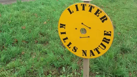 Mark Fishpool A round yellow sign with 'litter kills nature' message, placed on green grass verge.