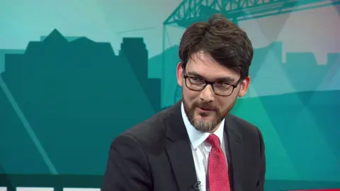 Markus Campbell-Savours appearing on BBC Politics North, wearing a black suit and bright red patterned tie. 