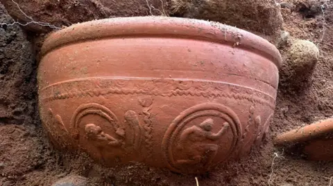Oxford Archaeology Large brown ceramic pot in brown dirt. It has patterns on it of people in circles.