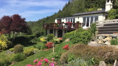 Aled Evans Photograph pf the remote bungalow in Maenan near Llanrwst, overlooking the Conwy Valley. The white building has a wooden patio perched above a sloping flower filled garden.