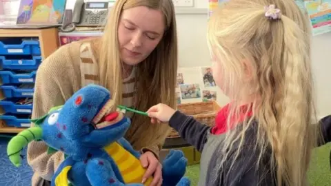 A young girl with blonde hair brushing the teeth of a blue toy dragon while a teacher looks on.