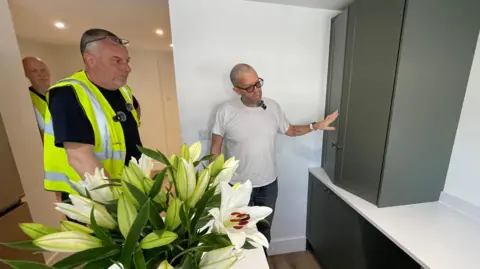 Stephen Huntley/BBC Three people are standing in a kitchen. Two on the left are wearing black T-shirts and Hi-Vis jackets. The other person, Jay House, is looking at one of the kitchen units on the right. 