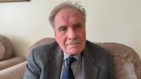 Francis Hillier sitting on a brown couch. He is wearing a grey suit and a dark blue tie.