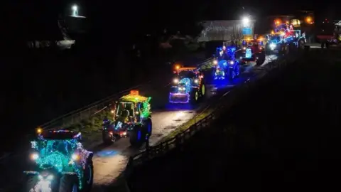 Joe Cullen Photography A line of illuminated tractors drive up a dark road. They are illuminated with loads of different coloured lights.