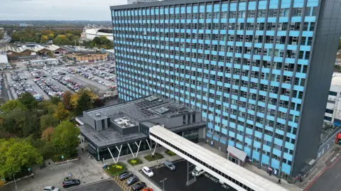 An aerial view of Hull Royal Infirmary - a large, modern hospital complex with a tall rectangular tower clad in alternating light blue and grey panels, featuring rows of identical windows. Below is a covered parking area with cars and vans parked within painted lines. Surrounding the building are parking areas filled with vehicles and tree‑lined streets. In the distance, trees and a stadium structure are visible under an overcast sky.