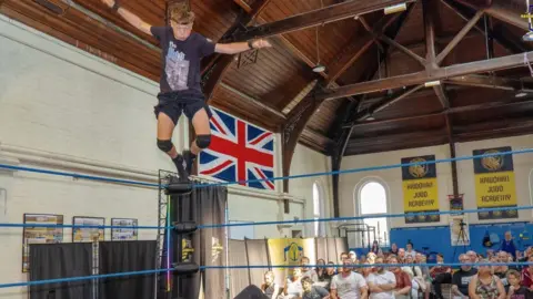 Rascals ProWrestling Academy Young wrestler in black kit jumps into a wrestling ring while club members look on.