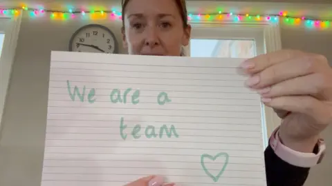 Vicki is holding a small piece of paper which says "We are a team" and then has a heart drawing in the corner and it is held in front of the camera. She is looking over the paper and is getting ready to move to the next one with her right hand. There are fairy lights on the wall behind her.