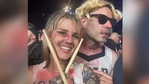 Supplied India Barton smiling holding two drumsticks either side of her face.