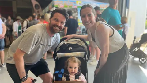Jack Maclean/BBC Ed and Rebecca Clark with their two-year-old daughter in a pushchair. They are all looking directly at the camera and smiling.