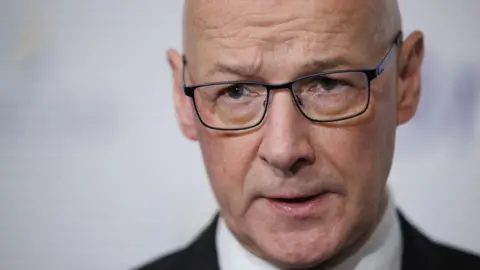 A bald man with glasses in a close-up shot. He is visible from the neck up and is wearing a dark suit and white shirt. 
