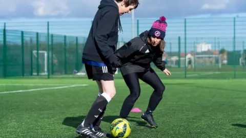A woman with short dark hair, dark sports top, black shorts, black socks and black football boots running after a yellow and black football with Clare Moss who has long brown hair, a blue and pink hat, green sports bottoms and black football boots. They are on a football pitch surrounded by a green cage.