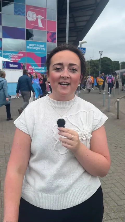A woman with a white short sleeve top on and brown hair tied back is standing facing the camera with a small mic in her hand.