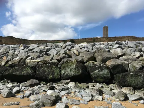 Andrew Woodger/BBC Crag Walk consists of rocks on the beach - with the sandy cliff behind them and the Naze Tower visible at the top of the cliffs on The Naze