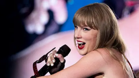 Getty Images Taylor Swift, a female singer holding a black microphone in her right hand singing into it while smiling.