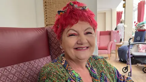 Darren Rozier/BBC A woman sits in a red armchair. She has bright red hair with matching red eyeshadow. She wears a vibrant green, purple and orange blouse. 
