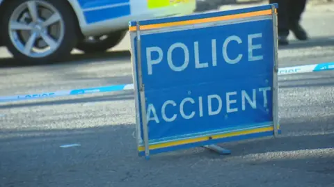 A blue Police Accident sign set up on the road. In the background there is a yellow and blue police car and cordon tape. 
