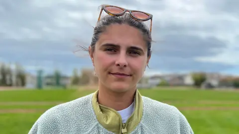 Claudia is standing on a wide grassy field under a cloudy sky. Low buildings and open green space extend into the distance, with sunglasses resting on the her head.