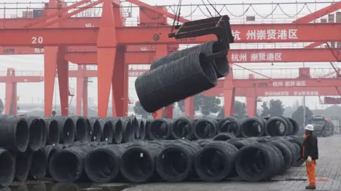 Getty Images A worker in orange pants, black jacket and a white helmet looks on at rolls of black steel being lifted by a red crane at a steel market in Hangzhou, China's eastern Zhejiang province on February 23, 2025. 