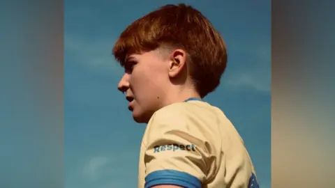 Ellie Scrivens A female footballer in her early 20s looks on during a match. Only her head and upper torso can be seen as it is a close-up shot of her side profile. She has short brown hair with buzzed sides and is wearing a yellow football jersey with blue arm cuffs. The word "respect" is printed on her sleeve in blue. Her mouth is slightly open and the sky behind her is slightly blue with very faint, wispy white clouds.