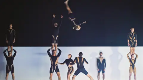 Circa tour video Three towers two of acrobats stand across the stage. On the far left and right, women stand with a partner on their shoulders, facing into the audience. In the centre, a third tower is mid-stunt, as a man on another man's shoulders throws a female acrobat into the air. She twists so her head is towards the ground mid-flight. Two others on the ground are waiting to catch her.