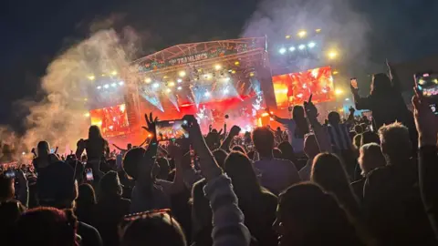A large crowd scene at night with hundreds of music fans facing a large stage lit by pink lighting