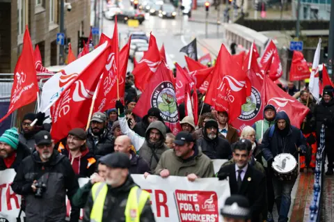 PA Media Striking workers marching along a Birmingham street. Some are holding a banner, others carrying red and white Unite the union flags