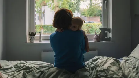 A stock image of a mother and child in a room, with a bed, a large window, with an E to the right. The mother is holding her child on one shoulder and she is wearing a blue top. 