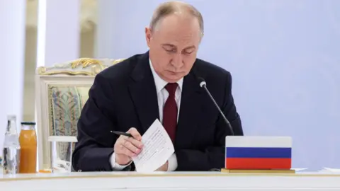 EPA Russian President Vladimir Putin attends the session of the Collective Security Council (CSC) of the Collective Security Treaty Organisation (CSTO) in Astana, Kazakhstan. Putin is holding a pen and a piece of paper, while in front of him sits a Russian flag and a microphone.