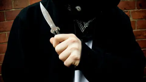The torso of a person wearing a black hooded jacket, a black and white bandana and a white T-shirt. They are holding a small knife in front of their body.