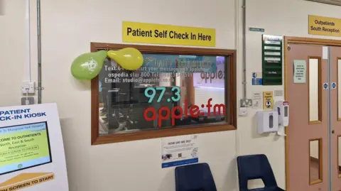 Apple FM Sign above a branded window saying 'Patient Self Check In Here'. The stickers on the window say 97.3fm apple:fm and there are two balloons tied to it. There are two dark plastic chairs under the window. There are double doors to the right of the image with a sign saying 'Outpatients South Reception' above them. There is a check in kiosk partly seen on the left of the image.