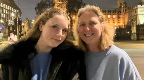 Louise Parry/BBC Zara has long fair hair and is wearing a black coat and blue top. She is standing next to her mother, who also has fair hair and is wearing a blue top. They are standing in front of the Houses of Parliament at night.