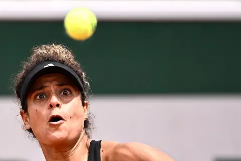 JULIEN DE ROSA / AFP / GETTY IMAGES A woman wearing a tennis visor stares intently at the ball as she prepares to strike it with her racket.