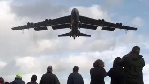 Grey US bomber plane photographed from below flying over a crowd of spectators near the RAF Fairford base 