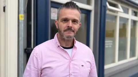 Un hombre con cabello oscuro mira a la cámara, lleva una camisa rosa.