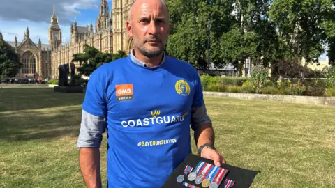 CRO Ian Bugler showing his medals during the demonstration outside the House of Commons. It is a sunny day.