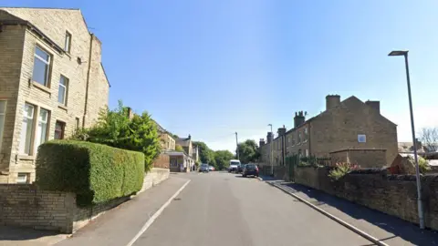 A row of houses either side of a road with a steep gradient. It's a clear day with blue sky. A house on the left has a neatly trimmed privet hedge. 