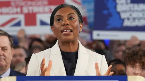 PA Media Kemi Badenoch wearing a white jacket and a black top standing in front of a crowd of people some of whom are holding up placards