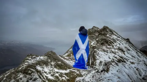 Cheryl Tracey A boy stands on top of a mountain with his back to the camera. He has a huge saltire flag around his shoulders which drapes to the ground.