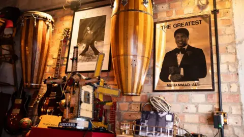 Jack Merriman Various guitars, drums and posters cover a brick wall in ambient lighting.