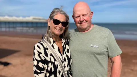 Tony and Liz Mabin stand together smiling at the camera with the beach, sea and pier stretched out behind them on a bright sunny day.
