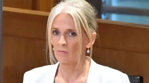 A woman with blonde hair, pinned up behind her head, wearing a white blazer. She is sitting on a chair in a room with wooden panels