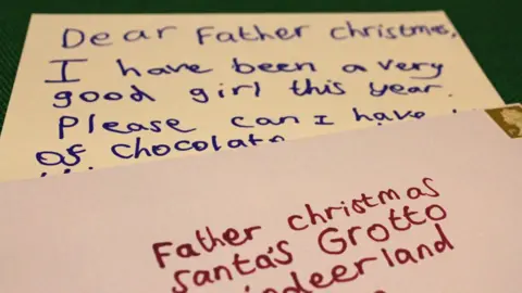 A handwritten letter, overlaid by its accompanying envelope. The letter is written to Father Christmas and the few words seen talk about being a good girl and asking for chocolate. The envelope is addressed to Father Christmas and addressed to the UK post office's official address for Christmas letters and is at least 10 years old