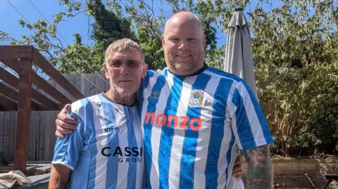 John is standing next to his son Scott. John is on the left and wearing a classic Coventry City shirt with the Cassidy Group sponsor logo. He as sunglasses on. Scott is wearing the 2026-27 Coventry City top with Monzo as the sponsor, is bald but with a stubble beard and tattoos on his left arm.