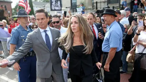 Karen Read is escorted through a large crowd of people holding signs outside a court house. She has long light brown hair and is wearing a black dress.